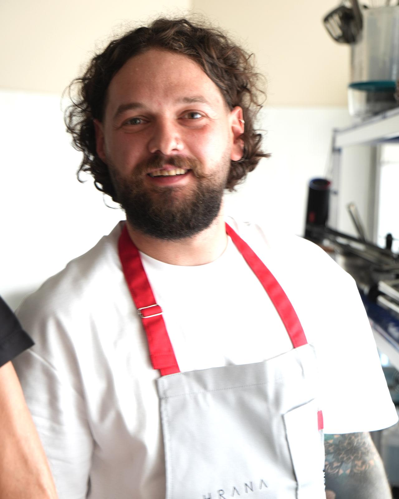 Chef Adrian Bașa, fondator și head chef HRANA, în șorț cu bretele roșii, zâmbind în bucătăria restaurantului